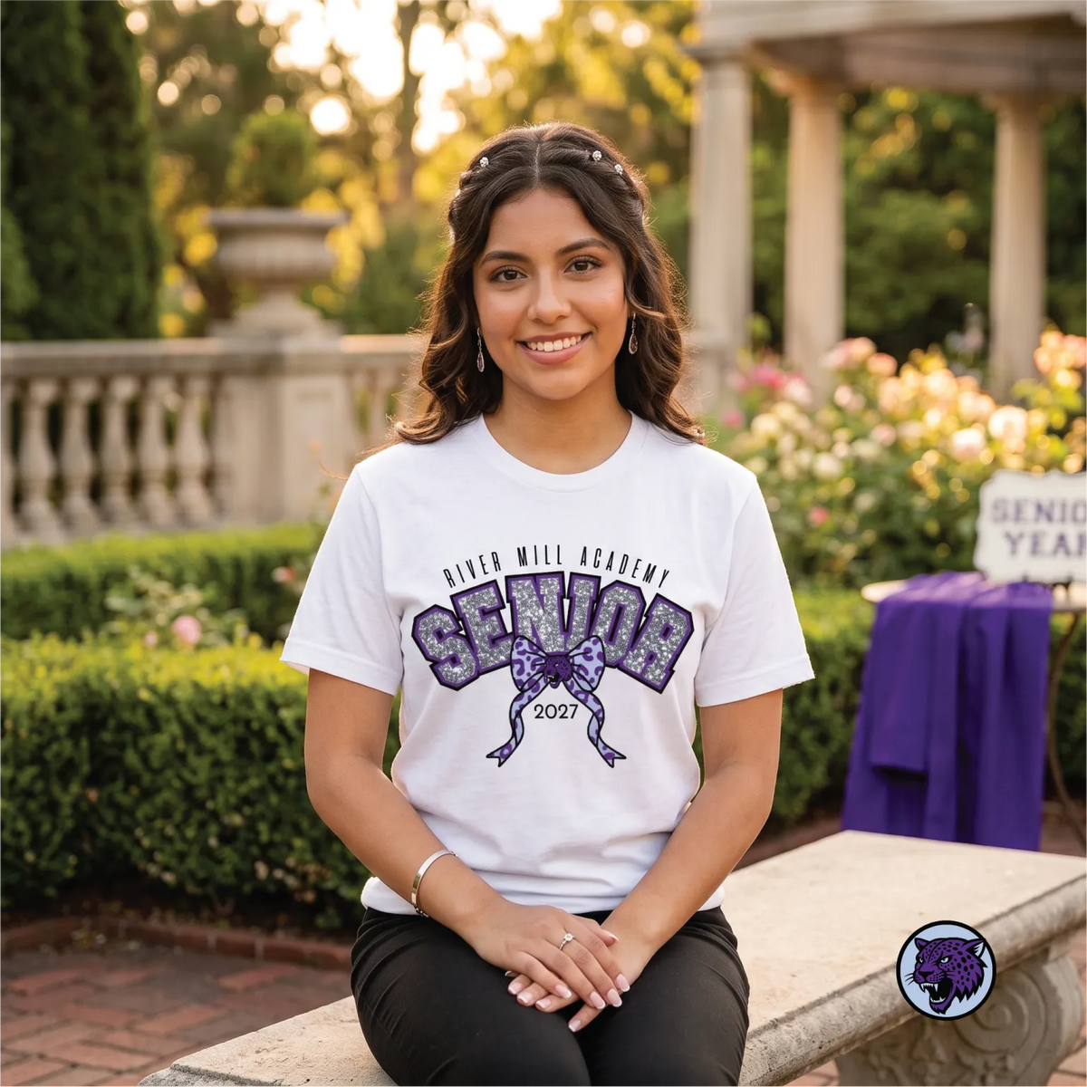 A smiling young woman sits on a stone bench outdoors wearing a white River Mill Academy 'Senior 2027' graphic t-shirt. The background shows a beautifully blurred garden with green hedges, flowers, and a stone colonnade.