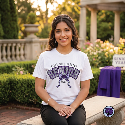 A smiling young woman sits on a stone bench outdoors wearing a white River Mill Academy 'Senior 2027' graphic t-shirt. The background shows a beautifully blurred garden with green hedges, flowers, and a stone colonnade.
